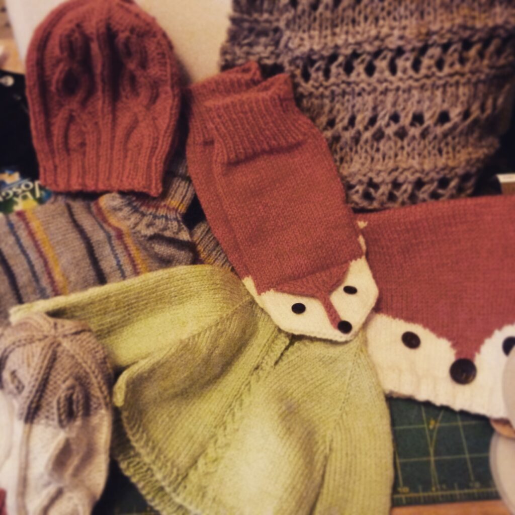 Photo regroupant plusieurs vêtements en tricot : bonnets et petit pull pour bébé, bonnet et mitaines à motif de renards, écharpe visible en arrière-plan et chaussettes épaisses pour chiller à la maison.